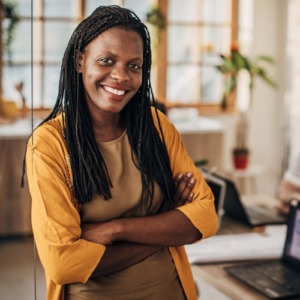 Young black businesswoman standing beside a desk in a sunny office with houseplants.